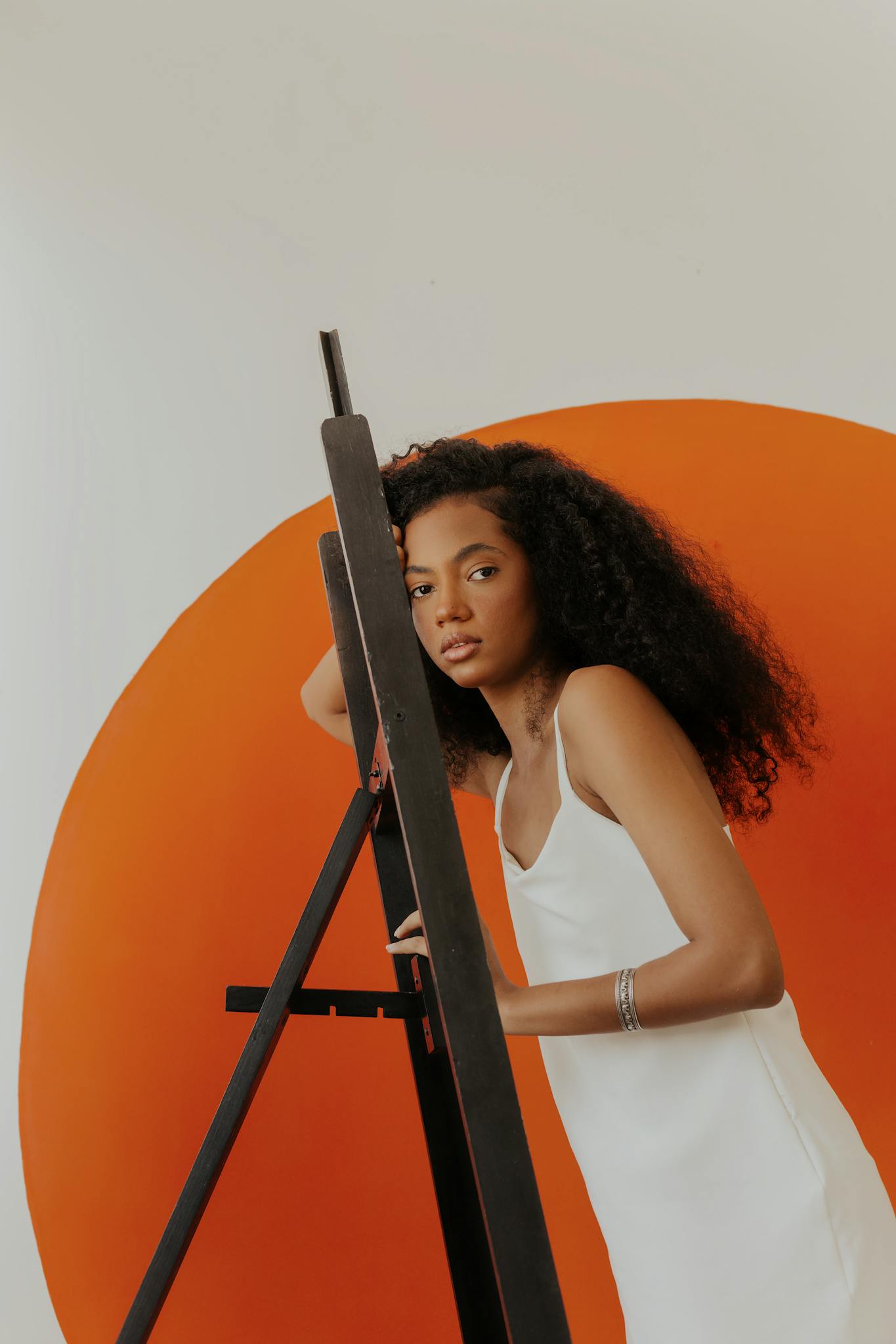 Artistic portrait of woman in white dress posing with an easel against vibrant orange background.